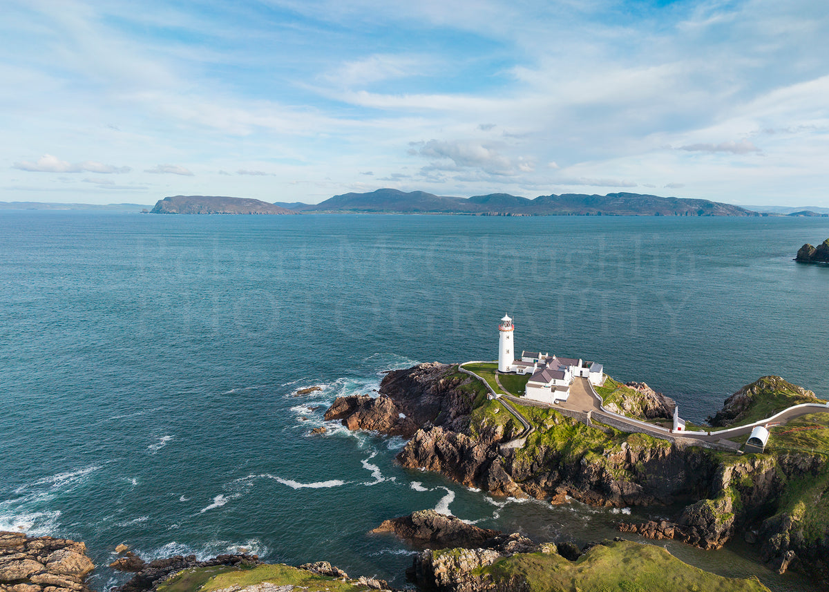 Aerial view of a white lighthouse complex on a rocky green cliff overlooking a deep blue ocean, with distant islands under a partly cloudy sky. A large semi-transparent watermark reading 'Robert McGlaughlin Photography' is visible across the center.