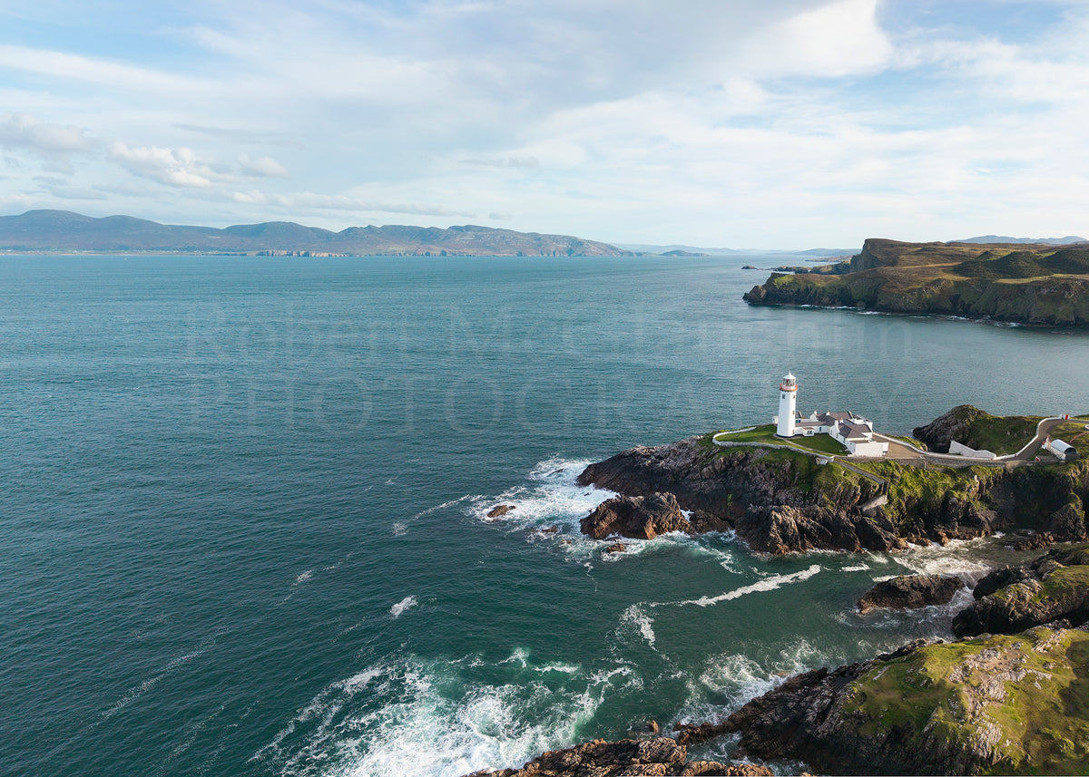 Aerial coastal landscape photograph showing a rocky shoreline with a white lighthouse on a grassy cliff, deep blue-green ocean water, distant mountains, and a cloudy sky. A faint 'Robert McGlaughlin Photography' watermark is visible across the centre.