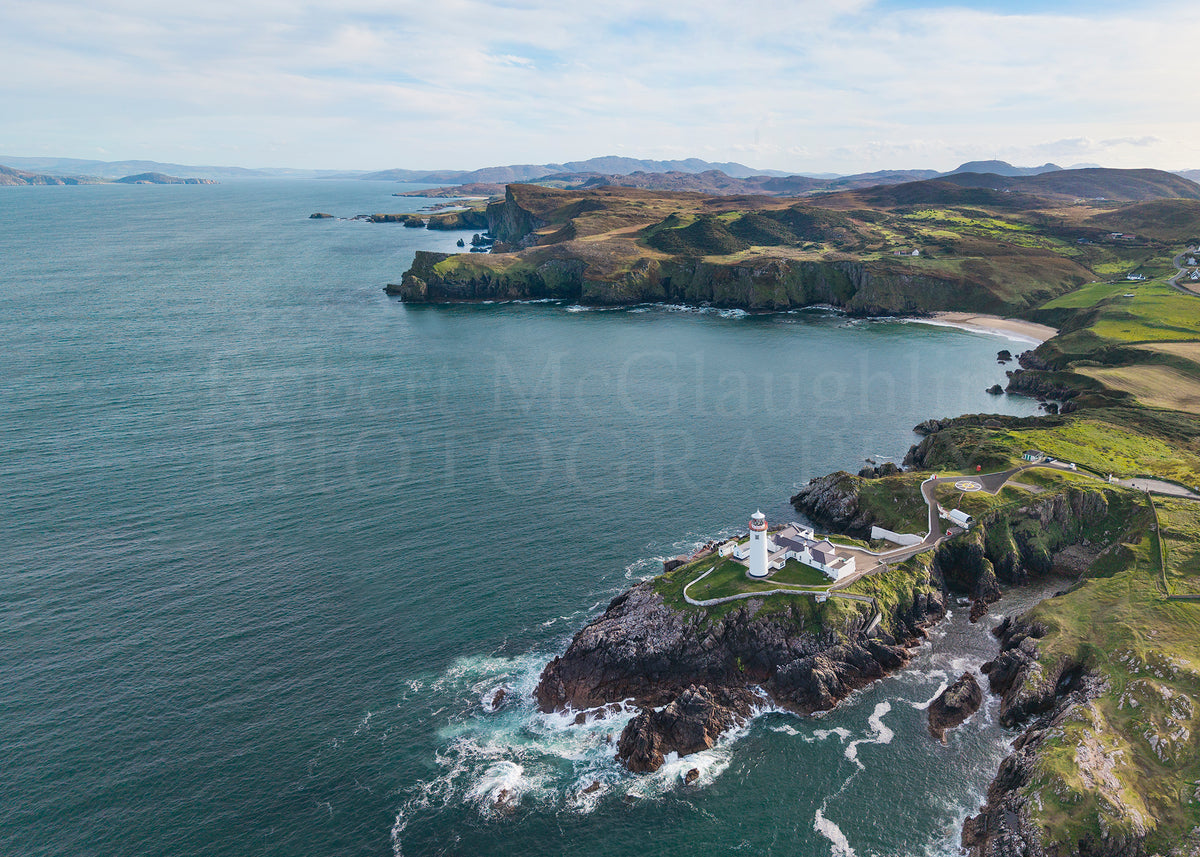 Aerial coastal landscape photograph showing rocky cliffs, green fields, a lighthouse, and deep blue ocean water under a cloudy sky. A faint 'Robert McGlaughlin Photography' watermark is visible across the center.